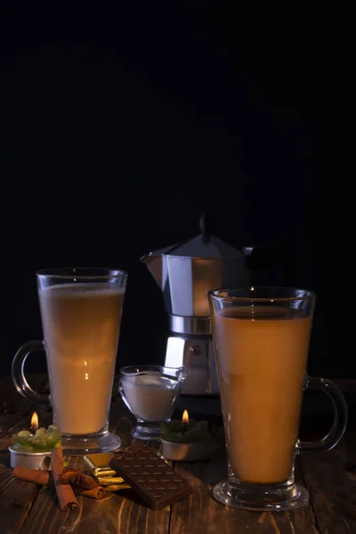 Two transparent glasses with latte, a coffee maker in the background on a wooden table with chocolate, anise, hazelnuts, lit by a candle flame. Black background.