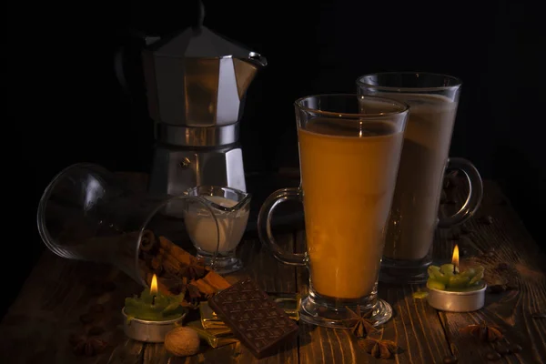 Dos vasos transparentes con café con leche, una cafetera en el fondo sobre una mesa de madera con chocolate, anís, avellanas, encendida por una llama de vela . — Foto de Stock