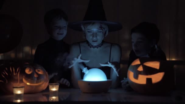 Mère avec deux fils jouant une sorcière à Halloween. Avec une lampe magique et une citrouille sur la table la nuit . — Video