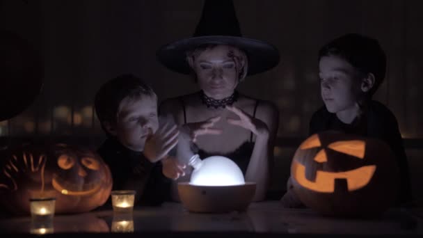 Mutter mit zwei Söhnen, die an Halloween eine Hexe spielt. mit einer Wunderlampe und einem Kürbis auf dem Tisch in der Nacht. — Stockvideo