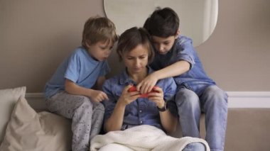 İki çocuklu beyaz bir kadın akıllı telefonu kanepede dinlenirken kullanırken eğleniyor. Mutlu çocuklar anneleriyle evde vakit geçirmeyi seviyor. Video oyunları