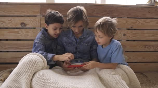 Mladá běloška se dvěma dětmi se baví pomocí smartphonu relaxaci na gauči, šťastní kluci tráví čas doma s mámou. Videohry — Stock video