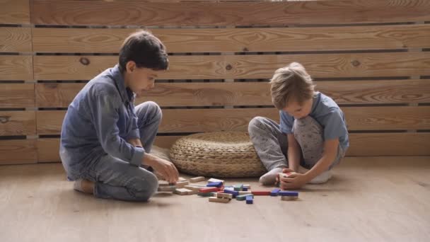 Dos hermanos chicos están construyendo una torre de bloques de madera sentados en el suelo — Vídeo de stock