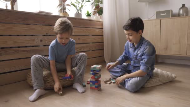 Два мальчика-брата строят башню из деревянных блоков, сидящих на полу — стоковое видео
