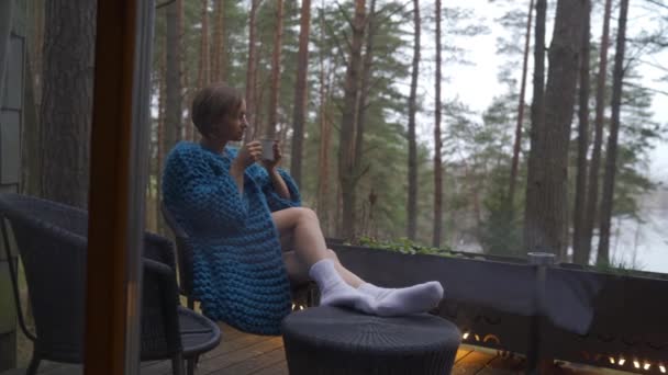 Attraktive Mädchen im Pyjama sitzt auf einem Balkon mit einer Tasse Tee oder Kaffee, genießt die Natur. — Stockvideo