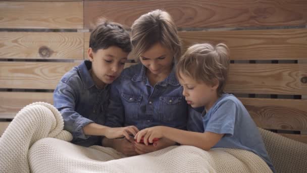 Jonge blanke vrouw met twee kinderen veel plezier met behulp van smartphone ontspannen op de bank, gelukkige jongens genieten van het doorbrengen van tijd thuis met mama. Videospellen — Stockvideo