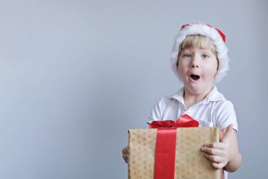 Elinde Noel hediyesi olan kırmızı şapkalı bir çocuk. Gri arka plandaki çocuk. Yeni yıl ve Noel konsepti.