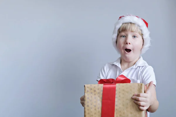 Kind met rode hoed met kerstcadeaudoos in de hand. Jongen op grijze achtergrond. Nieuwjaars- en kerstconcept. — Stockfoto