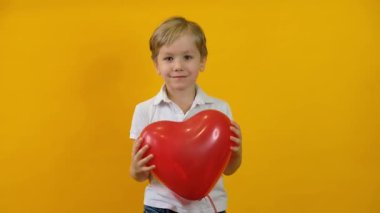 Kırmızı kalp şeklinde balon tutan ve sarı arka planda gülümseyen neşeli küçük çocuk.