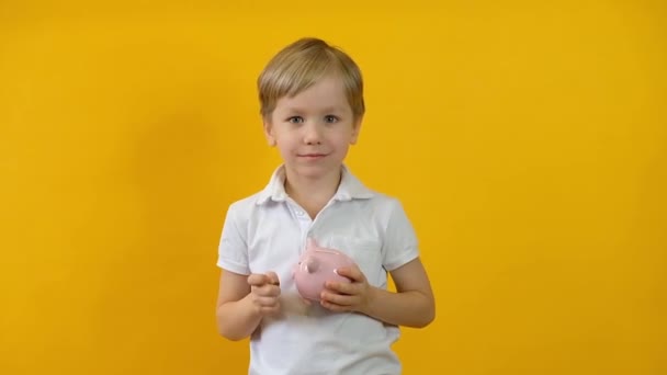 Loira criança menino colocando moeda para um porquinho banco isolado sobre fundo amarelo — Vídeo de Stock
