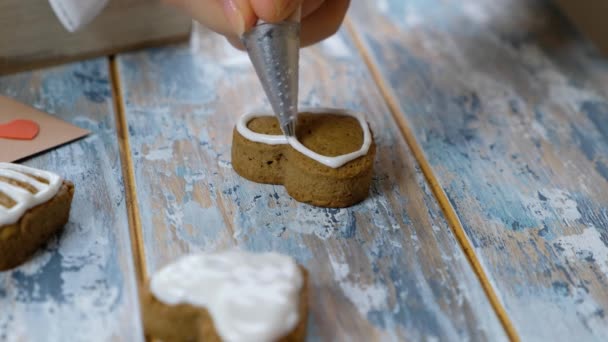 Mujer decorando galletas de jengibre en forma de corazón para el día de San Valentín o Navidad, cubriendo con esmalte blanco, de cerca — Vídeos de Stock