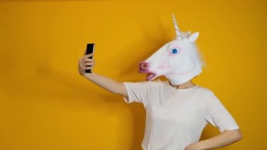 Strange funny video - woman with unicorn head making selfie with smartphone over yellow background