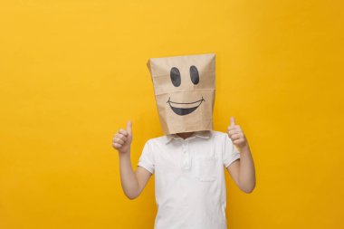 Kafasında kese kağıdıyla duran sevimli bir okul çocuğu. Sarı arka planda mutlu bir yüz, konsept.