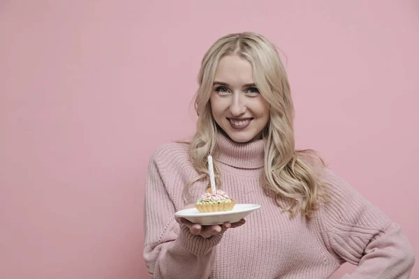 Happy blond woman in pink sweater holding cake with candles, celebrating birthday on pastel pink background. Copy space