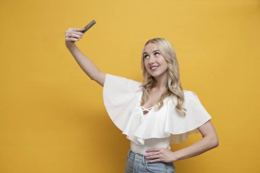 Beyaz bluzlu mutlu Avrupalı kadın sarı arka planda akıllı telefonuyla selfie çekiyor. Boşluğu kopyala