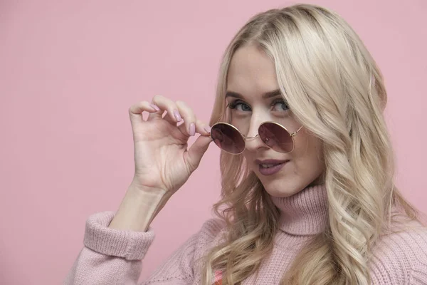 Happy coquettish woman face with blond hair in glasses and wool sweater on pastel pink background. Close up