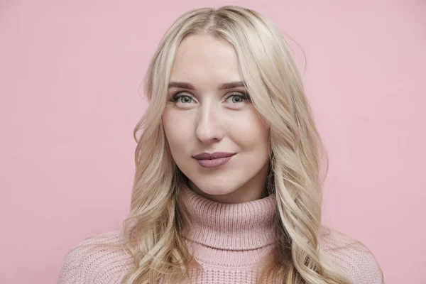 Sarı saçlı, yün kazaklı, pastel pembe arka planda kameraya bakan mutlu bir kadın yüzü. Kapat.