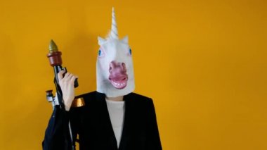Weird funny video - business woman in a suit with unicorn head holding a gun over yellow background