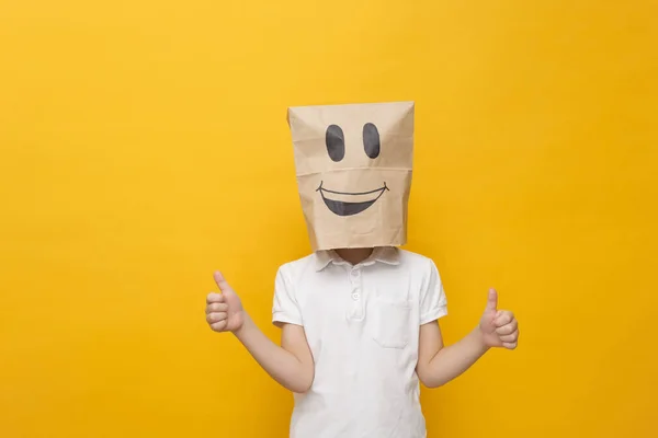 Lindo niño de la escuela de pie con una bolsa de papel en la cabeza - cara feliz en el fondo amarillo, concepto de emoción hppy — Foto de Stock