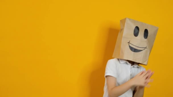 Kisfiú egy táskával a fején tapsol és táncol boldog mosolygós arccal sárga háttéren — Stock videók