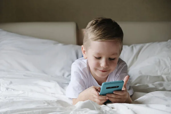 Lindo niño pequeño jugando juego móvil, acostado en una cama y sosteniendo el teléfono inteligente — Foto de Stock