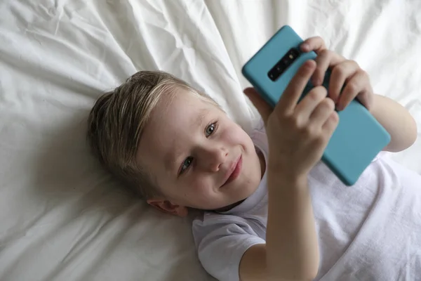 Lindo niño pequeño jugando juego móvil, acostado en una cama y sosteniendo el teléfono inteligente — Foto de Stock