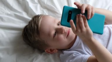 Küçük şirin bir çocuk mobil oyun oynuyor, yatakta uzanıyor ve akıllı telefon tutuyor. Üst görünüm
