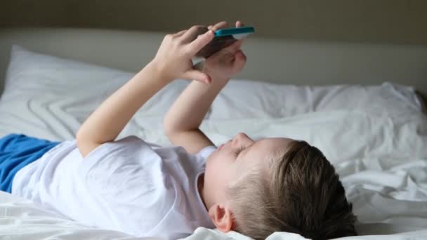 Criança menino bonito jogando jogo móvel, deitado em uma cama e segurando smartphone — Vídeo de Stock