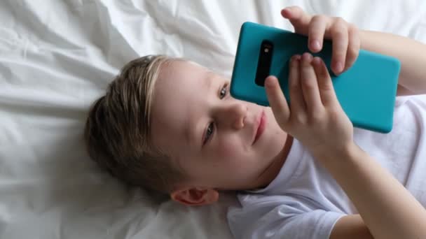 Criança menino bonito jogando jogo móvel, deitado em uma cama e segurando smartphone. Vista superior — Vídeo de Stock
