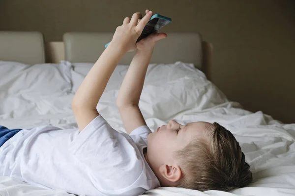 Söt liten pojke småbarn spelar mobilspel, ligger på en säng och håller smartphone — Stockfoto