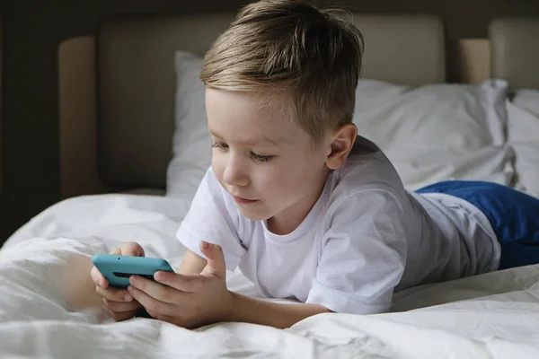 Lindo niño pequeño jugando juego móvil, acostado en una cama y sosteniendo el teléfono inteligente — Foto de Stock