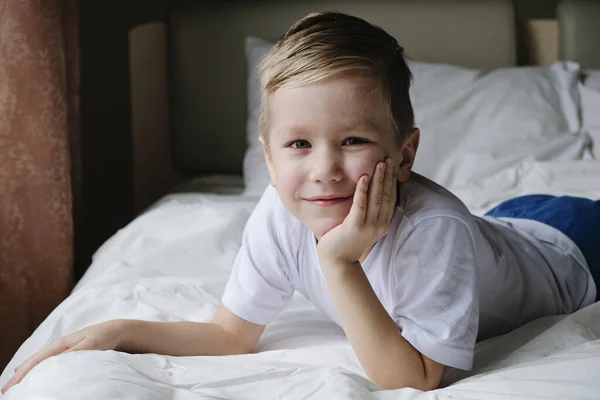 Schattige kleine jongen peuter liggend op wit bed, kijkend naar de camera en glimlachend. Ochtend levensstijl concept — Stockfoto