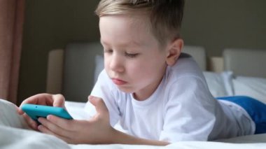 Sevimli küçük çocuk mobil oyun oynuyor, yatakta uzanıyor ve akıllı telefon tutuyor.
