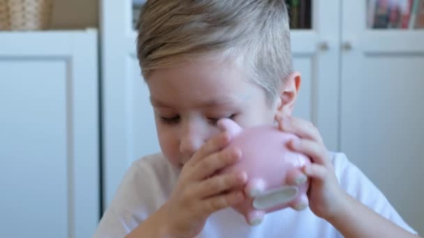 Schattig jongetje zet een munt in een spaarvarken bank, close-up — Stockvideo