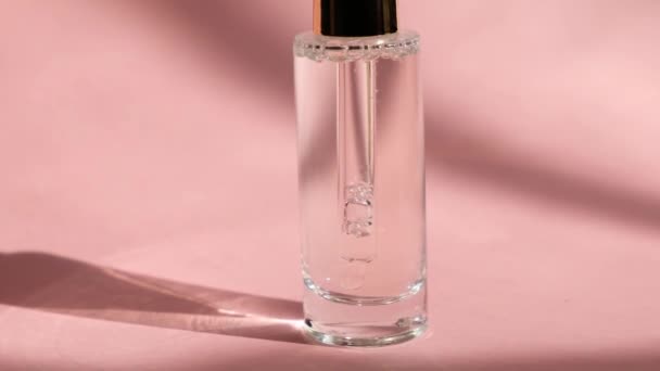 Сыворотка капельницы в прозрачной стеклянной бутылке на розовом фоне и женская рука, закрыть. Красота ухода за кожей, косметическая эссенция — стоковое видео