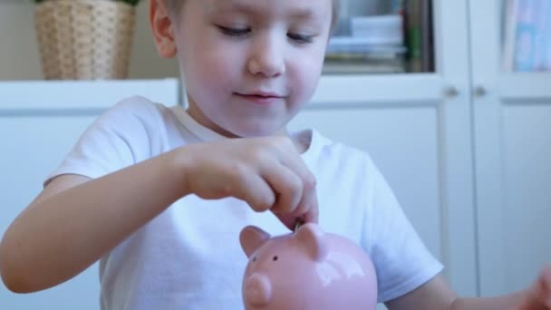 Bonito menino coloca uma moeda em um banco porquinho, de perto — Vídeo de Stock