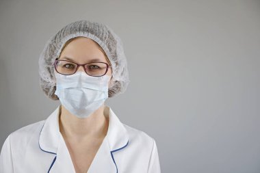 Tıp çalışanı portresi. Gri arka planda koruyucu maskeli ve gözlüklü kendine güvenen kadın doktor..