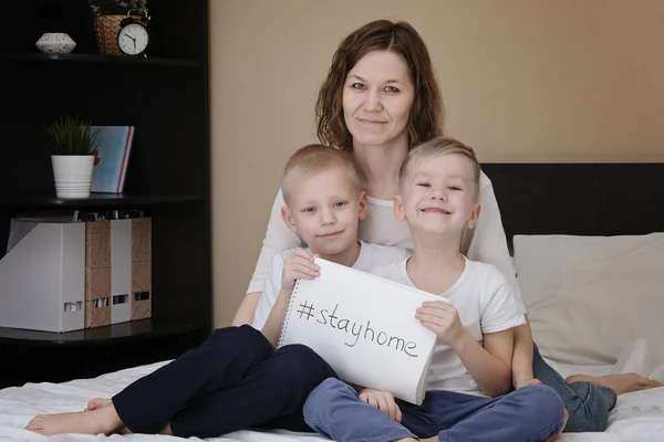 家人的母亲带着孩子坐在家里的床上，手里拿着一张写着"待在家里"的白纸。Coronavirus检疫 — 图库照片