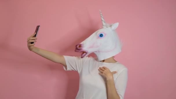 Странное смешное видео - женщина с головой единорога делает селфи со смартфоном на розовом фоне — стоковое видео