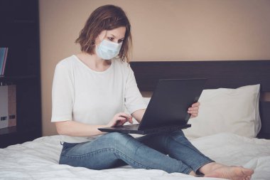 Maskeli bir kadın dizüstü bilgisayarla yatakta çalışıyor. Coronavirus karantinası, tecrit süresi - 19. Sosyal uzaklık