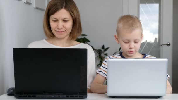 Uma jovem mulher e seu filho sentam-se em uma casa durante a quarentena e estudam na escola da internet usando um computador. Auto-isolamento — Vídeo de Stock