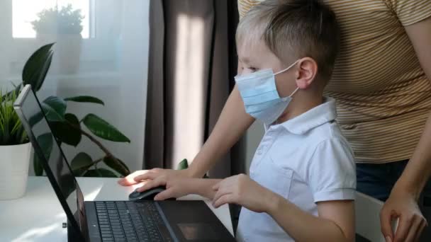 Дошкольный мальчик в медицинской маске обучение с помощью компьютера с мамой, концепция дистанционного образования — стоковое видео
