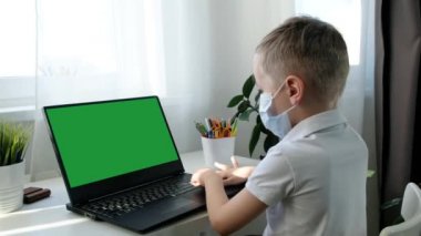 Tıp maskeli şirin bir okul çocuğu bilgisayar öğreniyor, uzaktan eğitim kavramı. Arkadan görüntüle, Yeşil Ekran