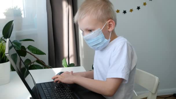 Милый школьник в медицинской маске, учится на компьютере, концепция дистанционного образования — стоковое видео