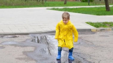 Sarı yağmurluk ve lastik çizmelerle su birikintisinde zıplayan komik çocuk.