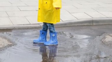 Sarı yağmurluk ve lastik çizme giyen komik çocuk su birikintisine atlıyor. Yaklaş.