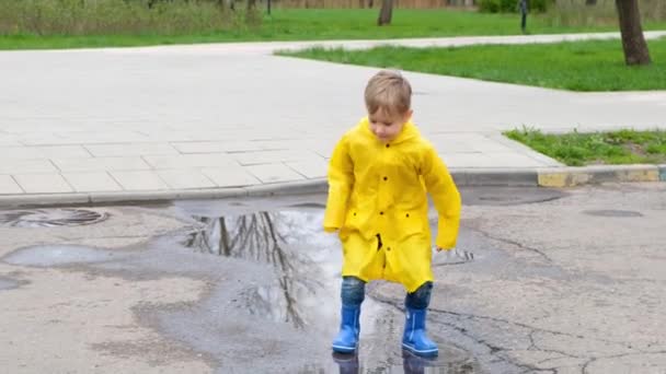 Vtipné dítě dítě ve žluté pláštěnce a gumové boty skákání v louži — Stock video