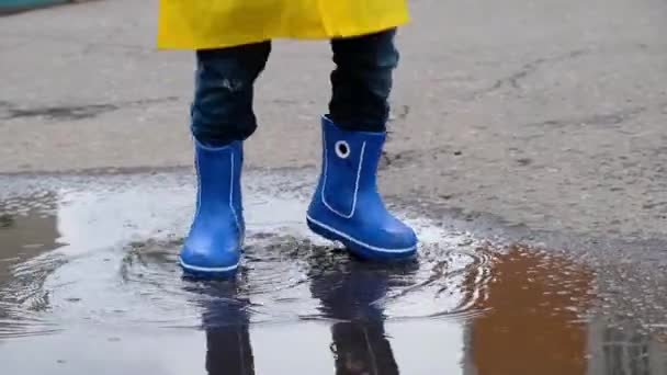 Vtipné dítě ve žlutém plášti a gumových botách skákající v kaluži, zblízka — Stock video