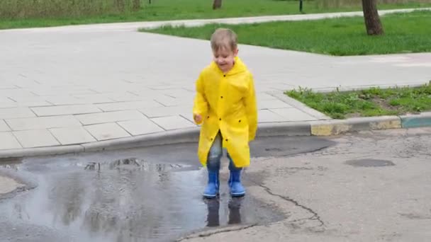 Αστείο παιδί με κίτρινο αδιάβροχο και λαστιχένιες μπότες να χοροπηδάει σε μια λακκούβα. — Αρχείο Βίντεο