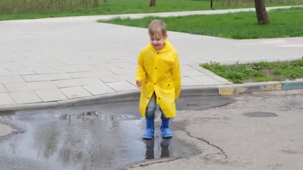 Funny child kid in a yellow raincoat and rubber boots jumping in a puddle — Stock Video